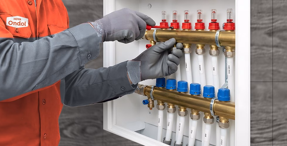 Plumber in a HOYA Ondol vest working on the floor heating manifold in a wall cabinet, with white pipe circuits running down to the floor.